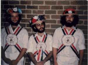 Three men with beards, straw hats with flowers, and blue, white and red baldricks.
