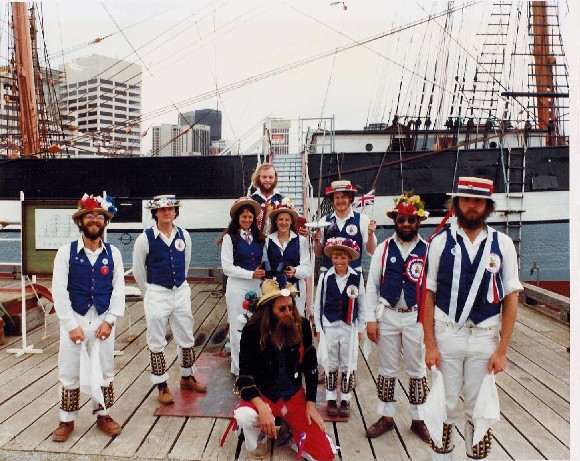 Dancers pose, smiling in front of a historic sailing ship.