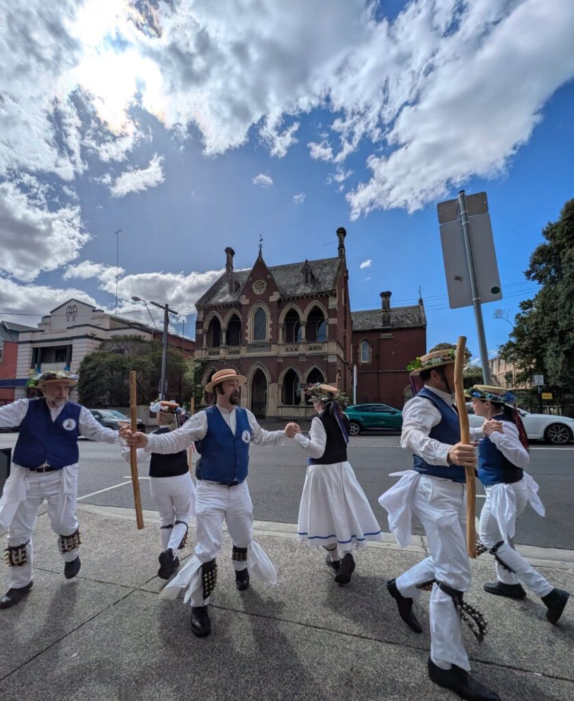 Dancers holding hands and long wooden sticks turn around in front of an old brick building with a bright sky above.