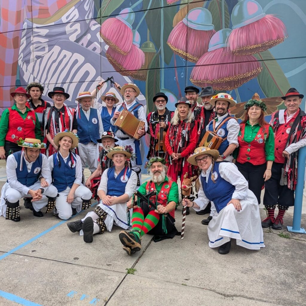 A combination of dancers from the different Melbourne Morris sides pose together under a bright mural.