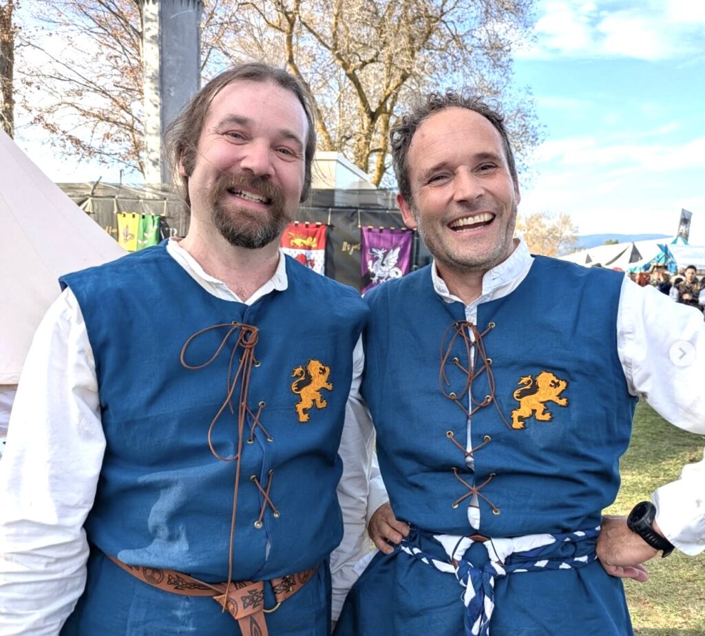 Justin and Keith smile at the camera, wearing the Britannia medieval kit of blue cottes over white shirts.